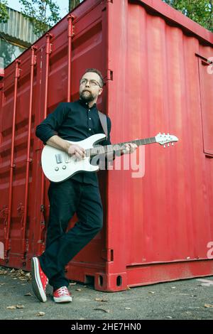 Uomo in nero con una barba che suona la chitarra elettrica, appoggiata al contenitore rosso in un ambiente urbano all'aperto in estate Foto Stock