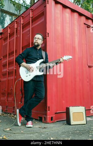 Uomo in nero con una barba che suona la chitarra elettrica con amplificatore combo, appoggiandosi al contenitore rosso sul luogo urbano all'aperto in estate Foto Stock