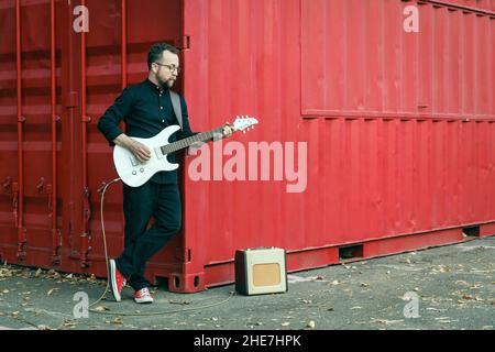 Uomo in nero con una barba che suona la chitarra elettrica con amplificatore combo, appoggiandosi al contenitore rosso sul luogo urbano all'aperto in estate Foto Stock