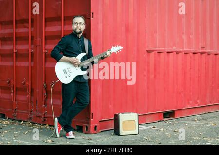 Uomo in nero con una barba che suona la chitarra elettrica con amplificatore combo, appoggiandosi al contenitore rosso sul luogo urbano all'aperto in estate Foto Stock