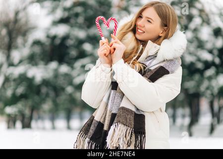 donna con cuore a forma di lecca, donna felice in abiti invernali, divertirsi con la caramella in mano all'aperto in una giornata nevosa all'aperto. Ragazza bionda con Foto Stock