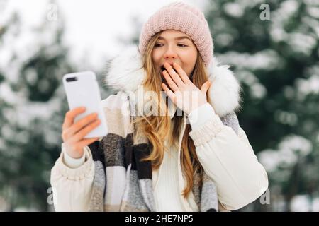 Immagine di una donna emozionata scioccata in un caldo cappello, usa uno smartphone e copre la bocca con la mano in sorpresa, guardando la macchina fotografica, una donna in wa Foto Stock