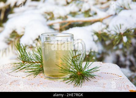 Infusione di tè con ago di pino caldo in bicchiere trasparente sul tavolo. Pino nevoso sullo sfondo, all'aperto nei freddi giorni invernali. Foto Stock