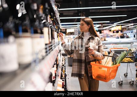 Giovane donna con carrello che guarda le scaffalature con vino in negozio. La ragazza sceglie il vino nel supermercato Foto Stock