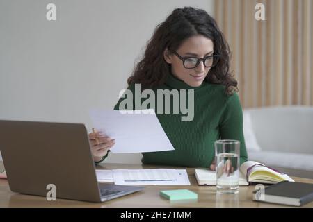 Giovane simpatica femmina italiana in occhiali preparazione report mentre si lavora su laptop in ufficio moderno Foto Stock