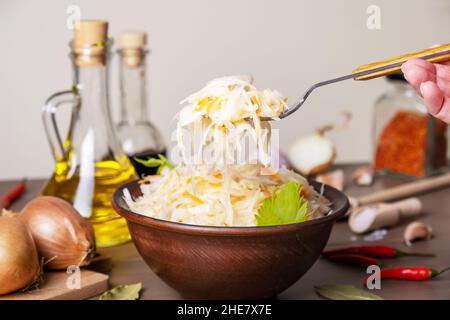 Cavolo fermentato in recipiente in ceramica sul tavolo con spezie e ingredienti. Porta a mano forchetta con insalata. Cibo sano, cucina tradizionale russa. Foto Stock