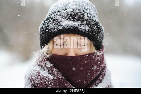Donna bionda in berretto beanie e naso coperto in piedi sciarpa con gli occhi chiusi nella nevicata invernale. Nordic inverno ritratto stagionale, tranquillo e calmo ou Foto Stock