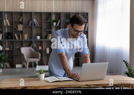 Uomo d'affari sorridente in occhiali utilizzando un computer portatile, in piedi alla scrivania Foto Stock