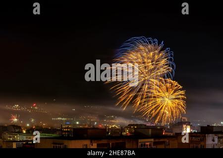 I fuochi d'artificio blu dorato brillano nel cielo scuro sulla città. La piccola città celebra l'anno nuovo con colorati pirotecnici di notte. Fuochi d'artificio per le feste. Foto Stock