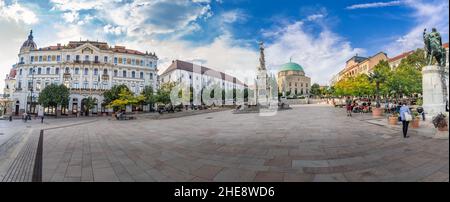 Vista panoramica della piazza Szechenyi con l'ex moschea trasformata in chiesa cattolica romana, la statua della trinità e altri edifici famosi nel centro di Pecs Foto Stock