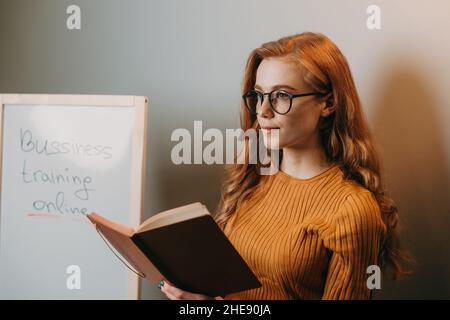 Insegnante con capelli rossi è in piedi e tenendo un libro di lezioni di fronte alla classe. Ritratto di donna incavato. Foto Stock