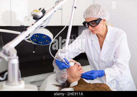 Cosmetologo che usa il laser per rifare la pelle del volto della donna matura Foto Stock