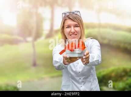 La ragazza tiene fuori la scatola del regalo con il nastro rosso all'aperto Foto Stock