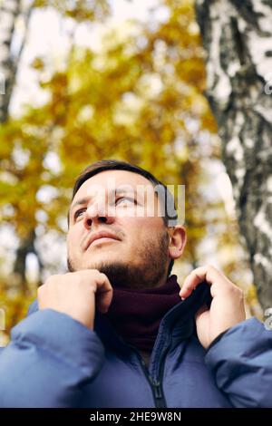 Ritratto di bell'uomo nella foresta autunnale in una giacca e sciarpa in giù. Vista frontale. Foto Stock
