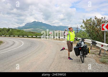 Una donna felice in bicicletta si trova vicino a una bici carica sull'autostrada tra le montagne. Foto Stock