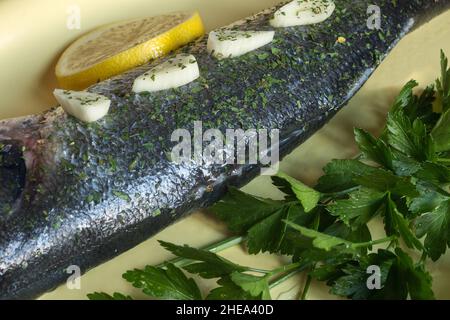 Rametti di prezzemolo fresco e specie secche e schiacciate su spigola cruda in un piatto di ceramica dipinta di giallo e un cuneo di limone. Cibo e pesce sani. Foto Stock