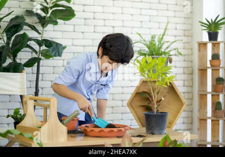 Il ragazzo asiatico trascorse le vacanze curando il giardino interno, mescolando il suolo e fertilizzante con la pala da giardinaggio blu. Foto Stock