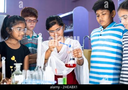 Panoramica dell'insegnante che spiega l'esperimento della scienza agli studenti con occhiali di protezione degli occhi al laboratorio di chimica - concetto di educazione Foto Stock