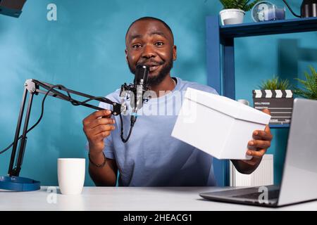 Sorridente influencer famoso parlare in microfono annunciando i vincitori dei social media in omaggio nella presentazione online live. Vlogger che detiene la confezione regalo per il vincitore del premio in vlogging studio. Foto Stock