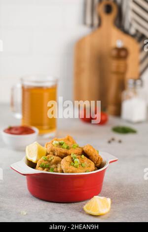 Pepite di pollo dorate in una ciotola rossa, cosparse di cipolle verdi e un cuneo di limone. Menu da pub Foto Stock