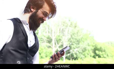 Uomo indù di successo con una barba e occhiali in un abito elegante con un telefono in mano all'aperto Foto Stock