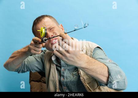 Divertente giovane uomo, pescatore con accessori di pesca seduta su sedia isolato su sfondo blu studio Foto Stock
