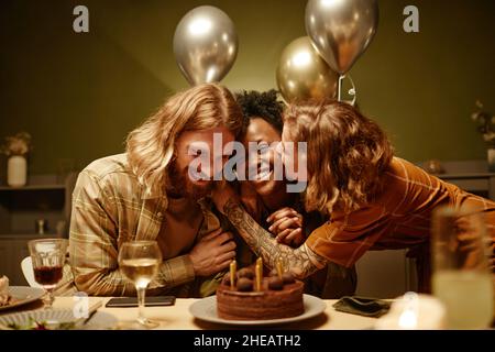 Giovane coppia felice congratulandosi con la loro amica con il compleanno, si siedono al tavolo baciandola e abbracciando Foto Stock