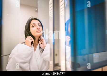 Il grazioso woamn le pennellerà i capelli e si guarda alla sua pelle chiara e luminosa nello specchio con luce di riflessione sullo sfondo Foto Stock