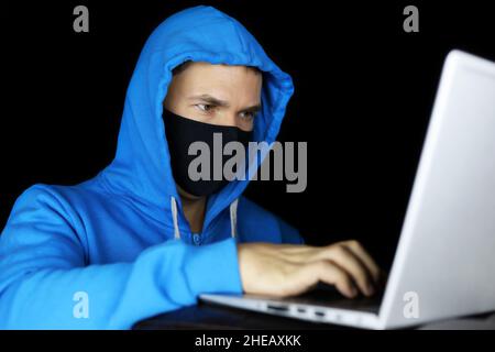 Uomo in maschera e felpa con cappuccio blu seduta con computer portatile su sfondo nero. Concetto di criminalità informatica, hacking e tecnologia Foto Stock