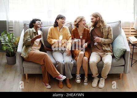 Gruppo di amici felici seduti sul divano bere champagne e ridere durante la conversazione in camera Foto Stock