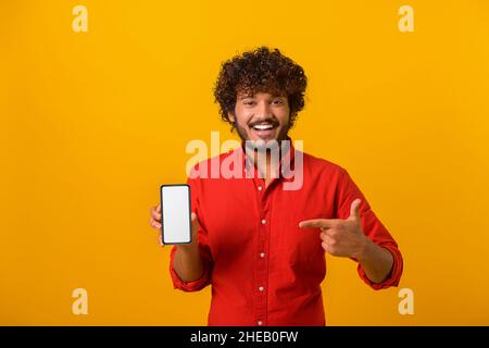 Uomo sicuro e positivo con la barba puntata verso lo smartphone con display bianco vuoto, che bragga con l'applicazione. Studio interno girato isolato su sfondo giallo Foto Stock