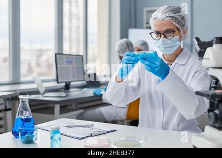 Giovane donna in maschera e guanti che tiene la piastra con i campioni mentre lavora sul posto di lavoro in laboratorio Foto Stock