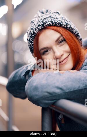 Foto all'aperto di notte di giovane bella ragazza sorridente felice godendo di decorazione festiva, in strada della città europea, indossando berretto di berlina lavorato a maglia Foto Stock