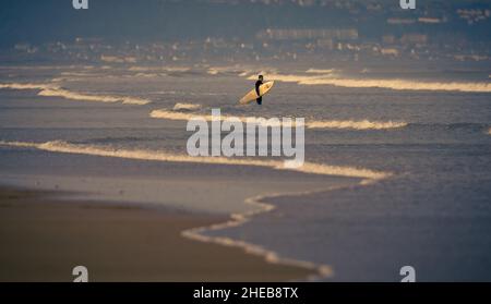 Un singolo surfer con bordo sotto il braccio aspetta in piedi nel surf guardando le onde su Saunton Sands Beach a nord del Devon. Foto Stock