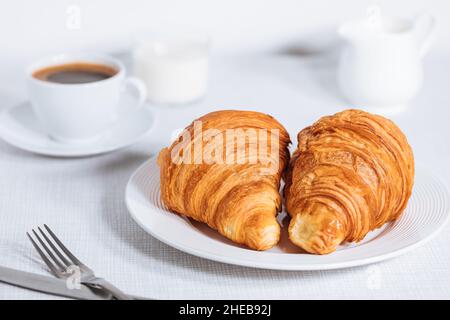 Croissant in un ambiente bianco luminoso, con una tazza di caffè e latte e altri croissant in uno sfondo sfocato. Ambiente colazione semplice ed elegante Foto Stock
