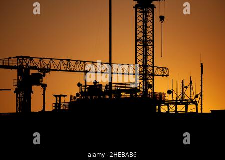 Cantiere al tramonto, gru e costruzione di una silhouette apicale contro il cielo. Foto Stock