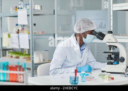 Giovane scienziato africano in maschera che esamina il campione di virus al microscopio mentre si siede al tavolo in laboratorio Foto Stock