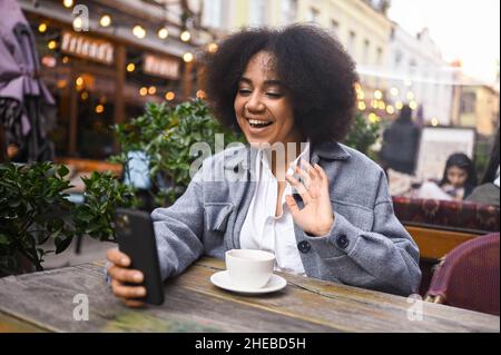 Ritratto Street style di attraente giovane felice afroamericana donna con capelli afro hanno video chat con smartphone all'aperto in caffè marciapiede. Eccitata sorridente femmina blogger trasmette live streaming Foto Stock