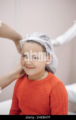 primo piano ritratto di sorridente bambina in maglione rosso su appuntamento medico, infermiere messo su cappuccio medico monouso Foto Stock