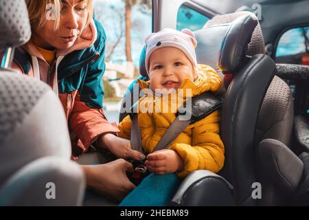 Ritratto di sorridente madre caucasica prepara il suo bambino adorabile per un viaggio in auto. Una donna allaccia le cinture di sicurezza in un seggiolino per bambini. Vista interna Foto Stock