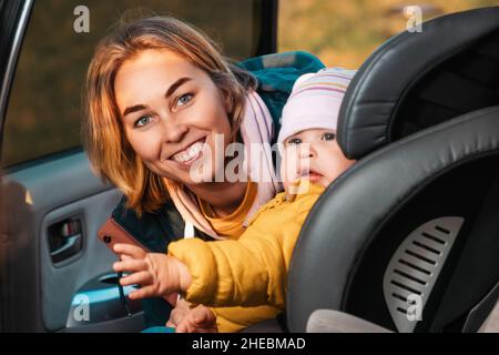 Ritratto di felice madre caucasica prepara il suo bambino carino per un viaggio in auto. Una donna allaccia le cinture di sicurezza in un seggiolino per bambini. Vista all'interno di un'auto Foto Stock