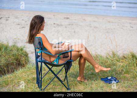 Donna, seduta su una poltrona da campeggio pieghevole, godendo una giornata nella natura in costume da bagno sulla spiaggia di Ersfjods a Senja, Norvegia settentrionale Foto Stock