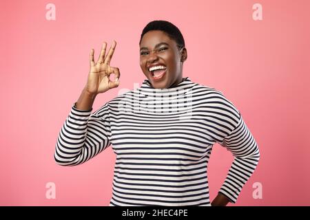 Joyful Overweight Black Lady gesturing OK in piedi su sfondo rosa Foto Stock