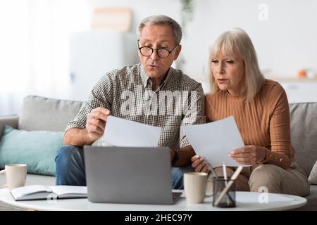 I coniugi senior leggono documenti e fatture seduti vicino a un computer portatile al chiuso Foto Stock