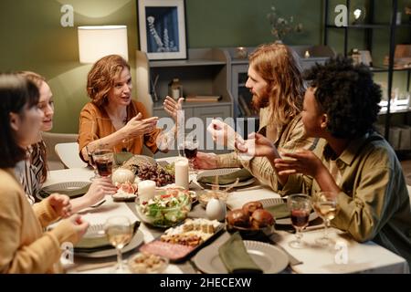 Giovane donna che parla con i suoi amici mentre mangiano a un tavolo festivo nel soggiorno Foto Stock