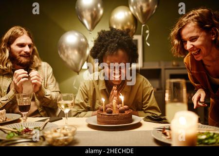 Donna felice africana seduta al tavolo con torta di compleanno con candele e ridere, che celebra il compleanno con gli amici a casa Foto Stock