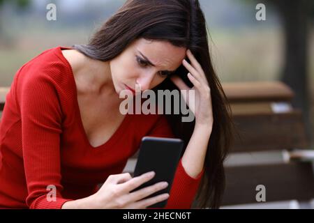 Donna preoccupata seduta su una panca che controlla il contenuto dello smartphone in un parco Foto Stock