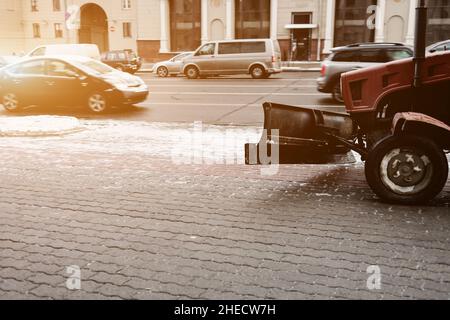Piccolo trattore con spazzaneve che guida su strada in città nella giornata invernale nevosa. Servizio comunale di pulizia della neve, trattore libera la strada da ghiaccio e neve. Foto Stock