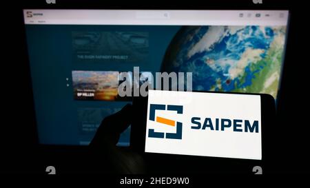 Persona che detiene smartphone con logo della società italiana Saipem S.p.A. di servizi petroliferi sullo schermo di fronte al sito. Mettere a fuoco sul display del telefono. Foto Stock