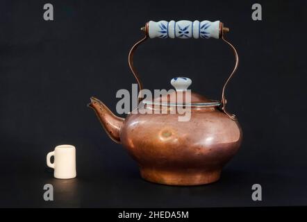 Teiera in rame antico o vintage, con una tazza di porcelano, davanti a uno sfondo nero. Foto Stock